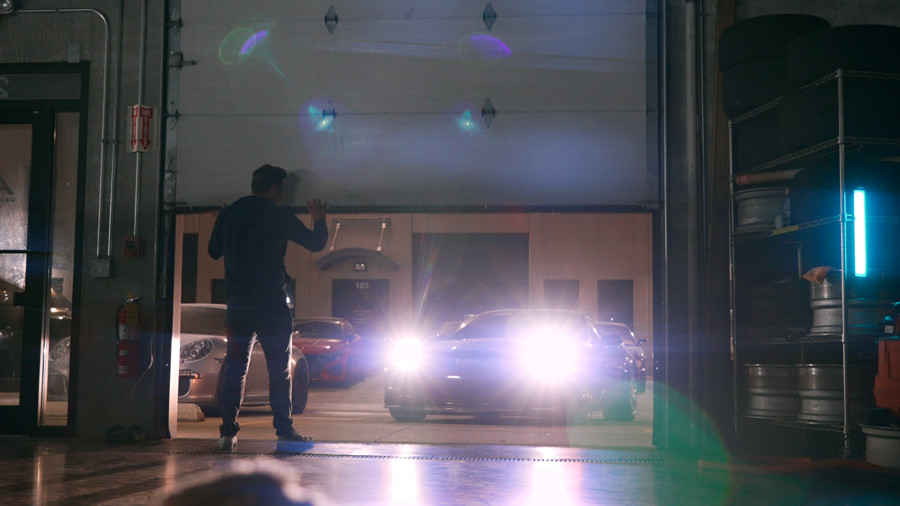 X standing in a garage beside bright headlights